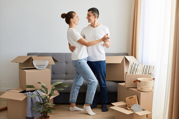 Fototapeta na wymiar Indoor shot of happy woman and man wearing jeans and white t shirts, couple posing among cardboard boxes and celebrating relocating in a new apartment, holding hands and dancing.
