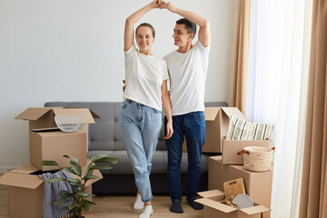 Fototapeta na wymiar Full length portrait of woman and man wearing jeans and white t shirts posing among carton parcels with belongings, standing near sofa and making roof with hands, moving in a new flat.