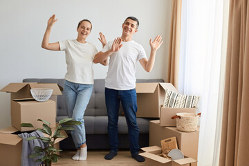 Fototapeta na wymiar Portrait of extremely happy woman and man wearing jeans and white t shirts posing among cardboard boxes and dancing, celebrating buying new their own apartment.