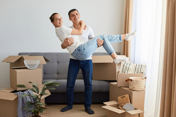 Fototapeta na wymiar Full length portrait of loving couple in a new house, woman and man wearing jeans and white t shirts posing among their personal piles in carton parcels, husband holding wife in hands.