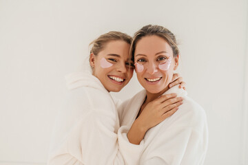 Positive two young caucasian women smile teeth standing inembrace on white background. Blondes wear bathrobes and patches during spa treatments. Concept of natural cosmetics, wrinkle smoothing - obrazy, fototapety, plakaty