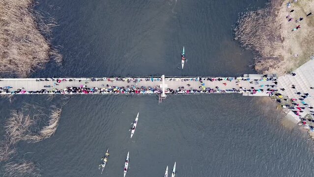 Top down video of boats canoes and kayaks going past the bridge. People standing on a bridge and looking. Colorful boats following the river. Marathon competition. Biathlon. Water sports. 
