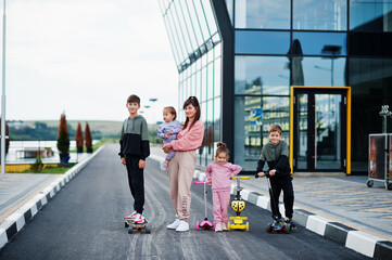 Young stylish mother with four kids outdoor. Sports family spend free time outdoors with scooters...