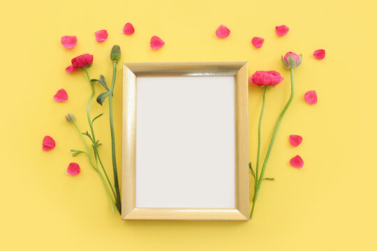 Top view image of pink buttercup flowers composition and empty gold photo frame over yellow pastel background