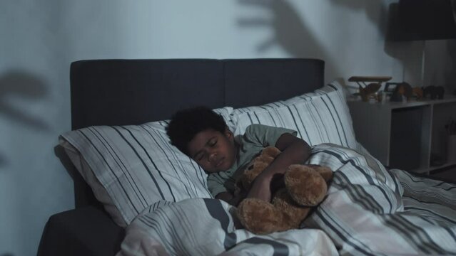 Medium of nine-year-old Black boy hugging toy bear, sleeping in his bed at home in dark, having nightmare of monsters reaching for him, waking up scared