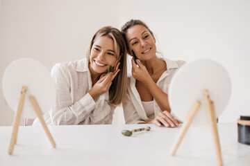 Happy young caucasian women use jade scraper while looking at mirror on white background. Blondes...