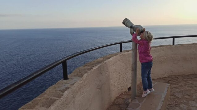 Little blonde girl having fun looking through a telescope on high elevated observatory desk. Wide shot