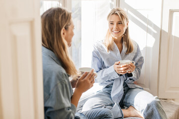 Smiling young caucasian women drink delicious coffee and gossip sitting on windowsill in morning....