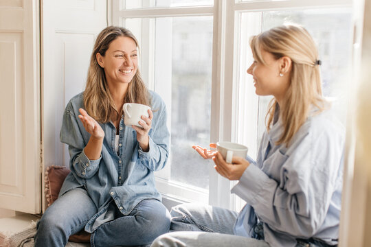 Two positive fair-skinned young women having cup enjoy strong coffee sitting on windowsill. Blondes in good mood wear casual clothes. Rest time concept.