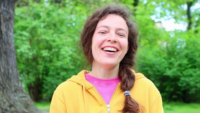 Cheerful casual young woman student laughing out loud at humorous joke looking at camera standing in green garden outdoors, with funny face enjoy sincere positive emotion, wearing yellow hoodie.