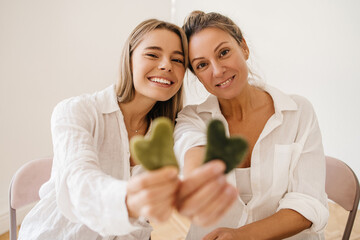 Cheerful young blonde caucasian ladies hold out gua sha scrapers to camera on white background....