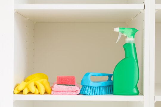 Brush, detergent bottle, rag, rubber gloves and sponge on white shelf inside opened wardrobe. Closeup. Set of colorful products for house cleaning. Front view. Home neatness.