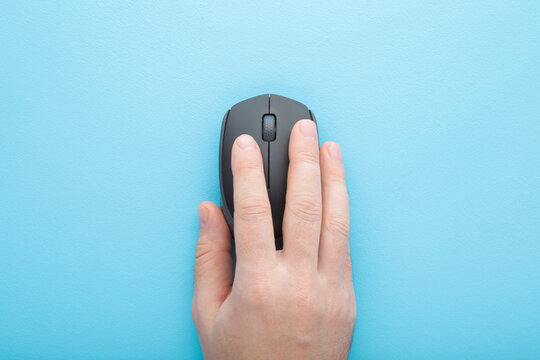 Young adult man hand using dark black computer optical mouse on light blue table background. Pastel color. Closeup. Top down view.