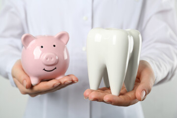 Dentist holding ceramic model of tooth and piggy bank on light background, closeup. Expensive...