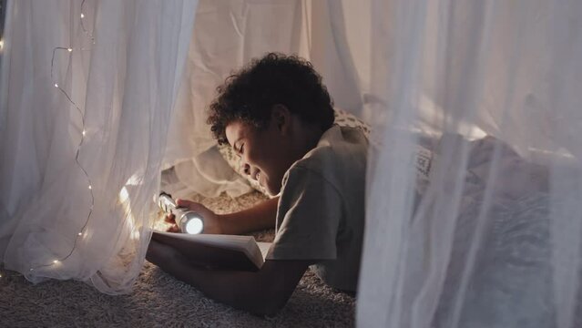 Medium side view of cute African American boy lying in dreamlike blanket fort, reading book using flashlight, smiling