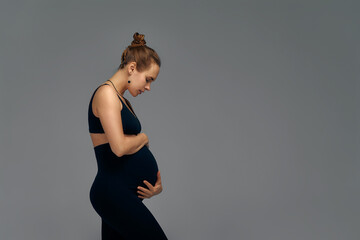 Cute pregnant belly isolated on gray. Side view of young pregnant woman embracing her abdomen with hands. Big belly on the third trimester of pregnancy close-up. Concept of pregnant life.