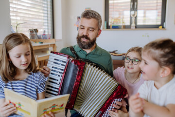 Cheerful father musician playing the accordion to his three little daughters at home. - obrazy, fototapety, plakaty