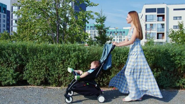 Pretty lady in sundress pushing baby stroller with sleeping toddler in front of her going at sidewalk. Stroll with child in pushchair at urban street. Young mom walking with son outside