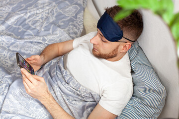 young man lying in bed in the morning wearing a sleep mask and checking social media