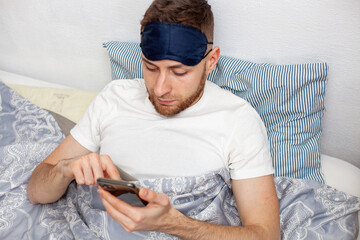 young man lying in bed in the morning wearing a sleep mask and checking social media