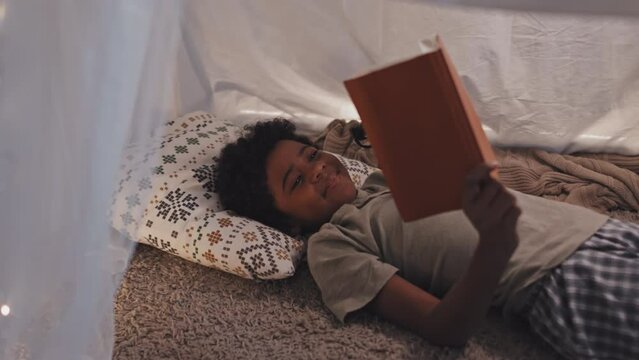 Medium of cute Black child lying on floor in handmade tent at home, reading book using flashlight at night, smiling