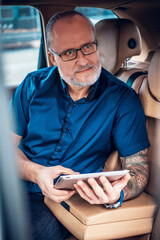 Fototapeta Portrait of old man with tattoo and glasses using tablet sitting inside of car in city. obraz
