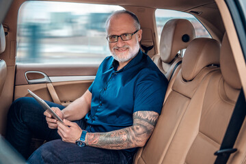 Fototapeta Portrait of tattooed glad old man executive with tablet sitting inside of car. obraz