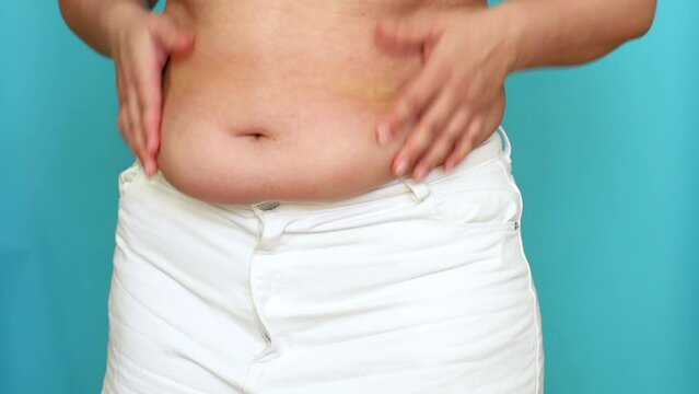 Unrecognizable fat plump overweight woman wearing white jeans, shaking belly with hands, squeezing excess flabby stomach on blue background. Body positive, massage, obesity, weight loss, liposuction.