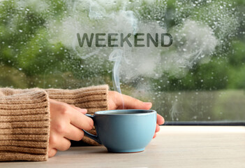 Happy Weekend. Woman with cup of hot drink at wooden table near window on rainy day, closeup