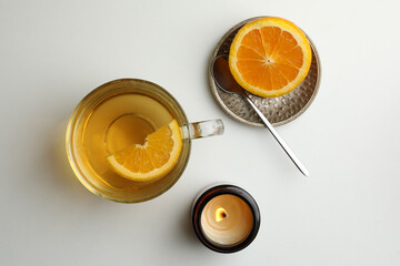 Cup of freshly brewed tea, burning candle and orange on light table, flat lay