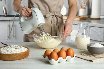 Woman whipping white cream with mixer at light grey table in kitchen, closeup