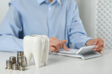 Woman using calculator at white table indoors, focus on ceramic model of tooth and coins. Expensive...