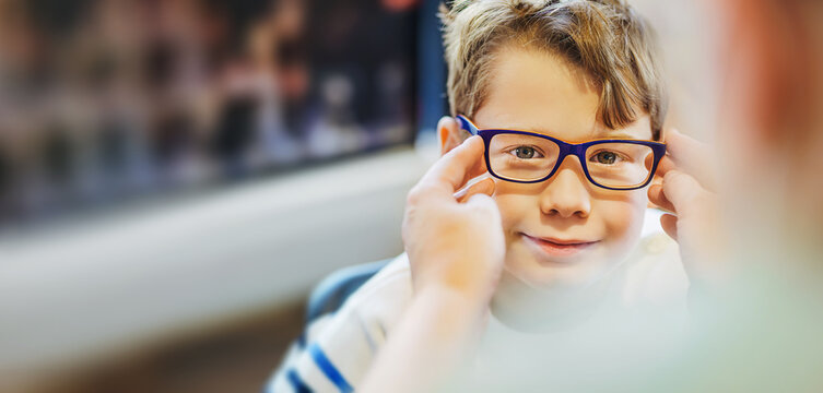  ophthalmologist trying eyeglasses on smiling boy in optics store, banner.
