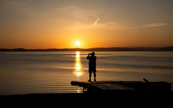 Silhouette of unknown person standing on beach platform with camera taking a picture of beautiful evening sunset.