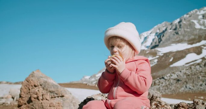 Cute toddler girl on travel with family sit on rock taking a break and eat green apple on epic mountain view. Concept of wanderlust lifestyle