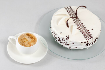 A cup of coffee and a white cake decorated with chocolate decor on a glass plate on a gray background. Selective focus.