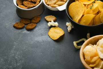 Unhealthy food. Snacks. All classic potato snacks with peanuts, popcorn and onion rings and salted pretzels in bowl plates on black concrete background. Unhealthy food for figure, heart, skin, teeth