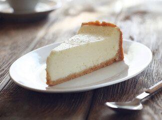 A piece of cheesecake cake lies in a plate on a wooden table. Behind is a cup. There is a spoon next to it.