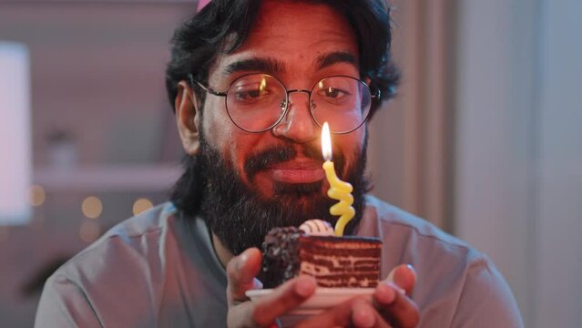 Portrait of multiracial man with thick black beard in glasses and festive hat with cake in light in evening indoors in dark says wish dream happily celebrates birthday blowing out burning candle