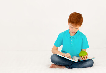 A cute red-haired boy is reading a book on the bed and holding a green apple in his hand