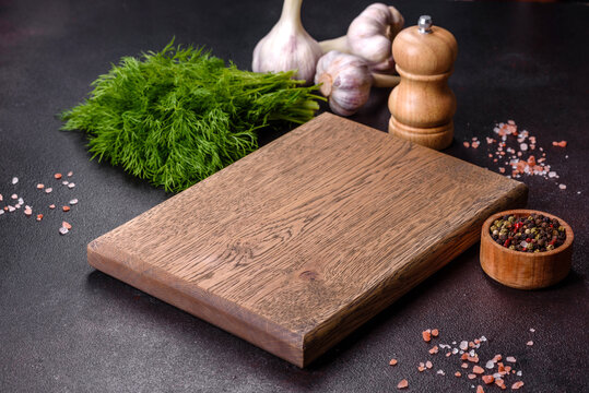 A wooden cutting board with spices, herbs, cherry tomatoes and salt on a black concrete background