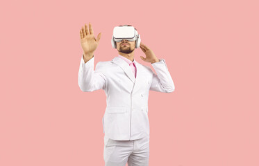 Man in white suit and VR headset standing against solid pastel pink background. Studio shot of young guy wearing VR glasses, experiencing virtual reality, exploring new world, touching virtual objects
