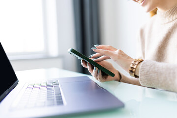 Close up of young woman hands using laptop checking smart phone at home