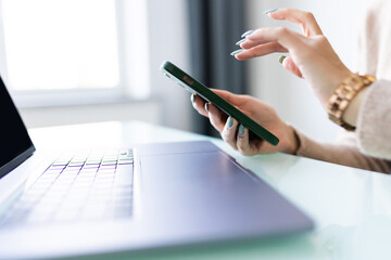 Close up of young woman hands using laptop checking smart phone at home