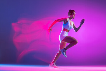 Obraz na płótnie Canvas Professional female athlete, runner training isolated on blue studio background in mixed pink neon light. Healthy lifestyle, sport, motion and action concept.