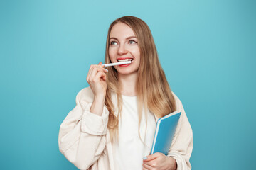 blonde girl student thoughtful, daydreaming and bit her handle with her teeth isolated on a blue studio background. Education concept