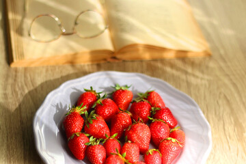 Lilac plate full of fresh strawberries, open book and reading glasses on wooden table. Selective focus.