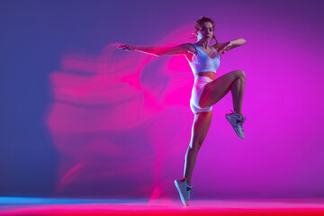 Fototapeta na wymiar Young female athlete, runner training isolated on blue studio background in mixed pink neon light. Healthy lifestyle, sport, motion and action concept.