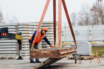 Fototapeta na wymiar Slinger in helmet and vest controls unloading of metal structures on construction site. White handyman unloads load.