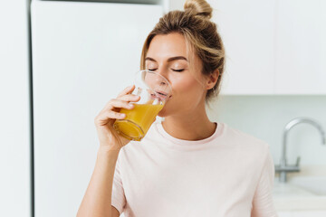 Woman drinking freshly squeezed homemade orange juice in white kitchen - obrazy, fototapety, plakaty